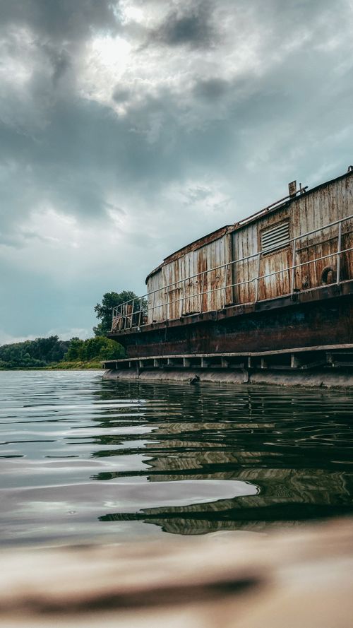 Free A weathered barge by a serene river with a dramatic cloudy sky, hinting at a storm. Stock Photo