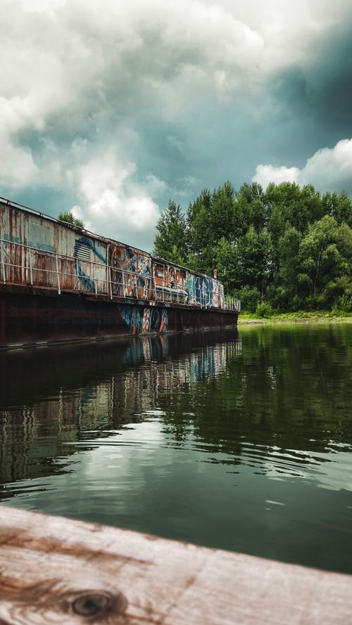 Free A serene view of a graffiti-covered bridge reflecting in a river under a cloudy sky. Stock Photo