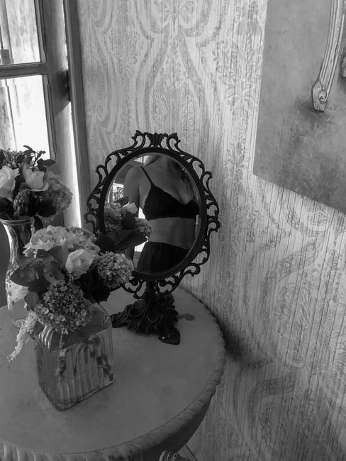 A black and white photo of a woman reflected in an ornate mirror, surrounded by flowers.