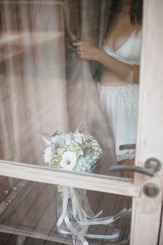 Elegant bridal bouquet and woman in lingerie through window curtain.