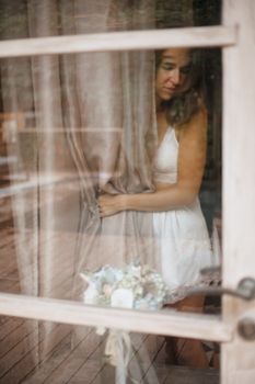 A woman in a white dress stands by a window holding curtains, reflecting introspection.