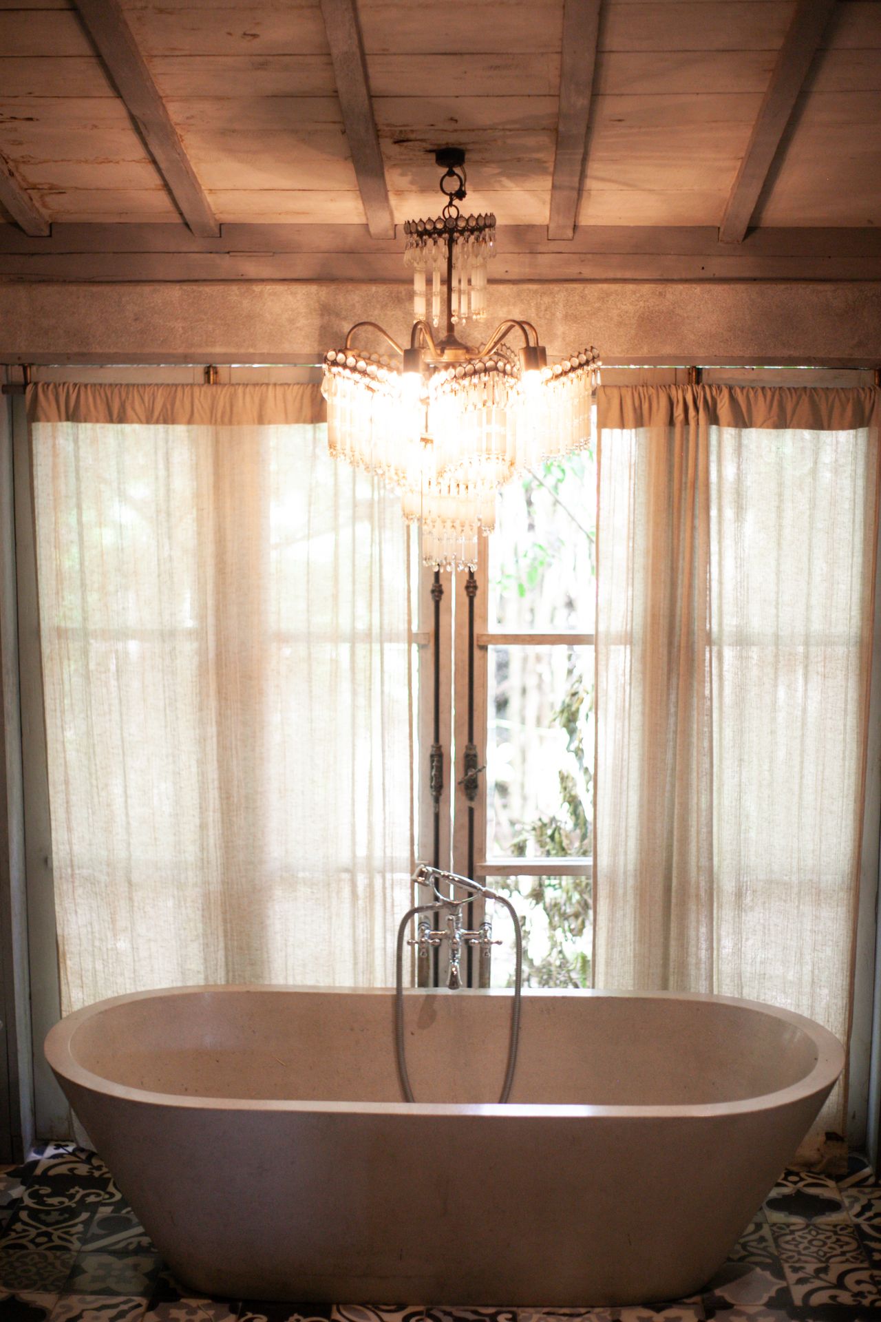 Crystal chandelier hanging over a freestanding stone bathtub in a bright bathroom with sheer curtains