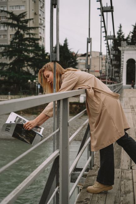 A woman leaning over a railing on a footbridge reading a magazine in an urban setting.