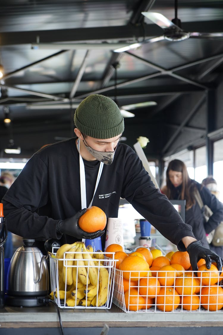 Man In Black Long Sleeve Shirt Wearing Face Mask Putting Oranges On Fruit Crate