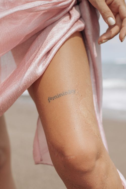 Free An artistic close-up of a woman's leg displaying a 'femininity' tattoo, captured on a sandy beach. Stock Photo