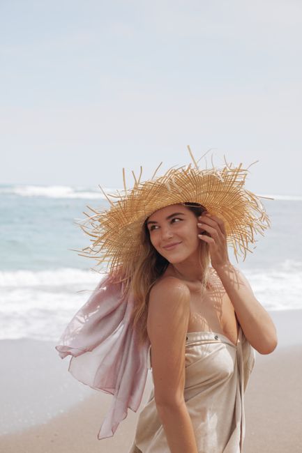 A smiling woman in a straw hat enjoys the sun at a beach, embodying summer relaxation.