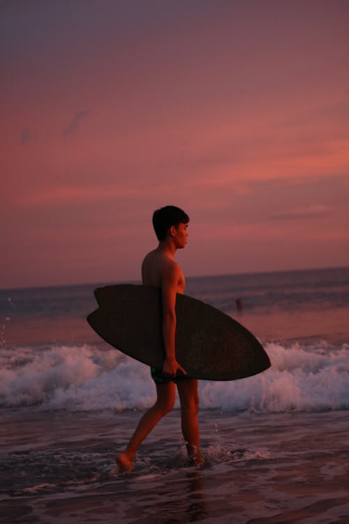 Free A Man Carrying a Surfboard Walking on the Seashore during Sunset Stock Photo