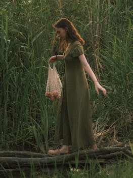A woman in a long green dress walks through lush greenery carrying a mesh bag filled with fruits.