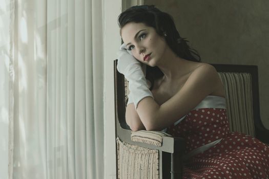 Elegant woman in a red polka dot dress sits thoughtfully indoors in a retro setting.