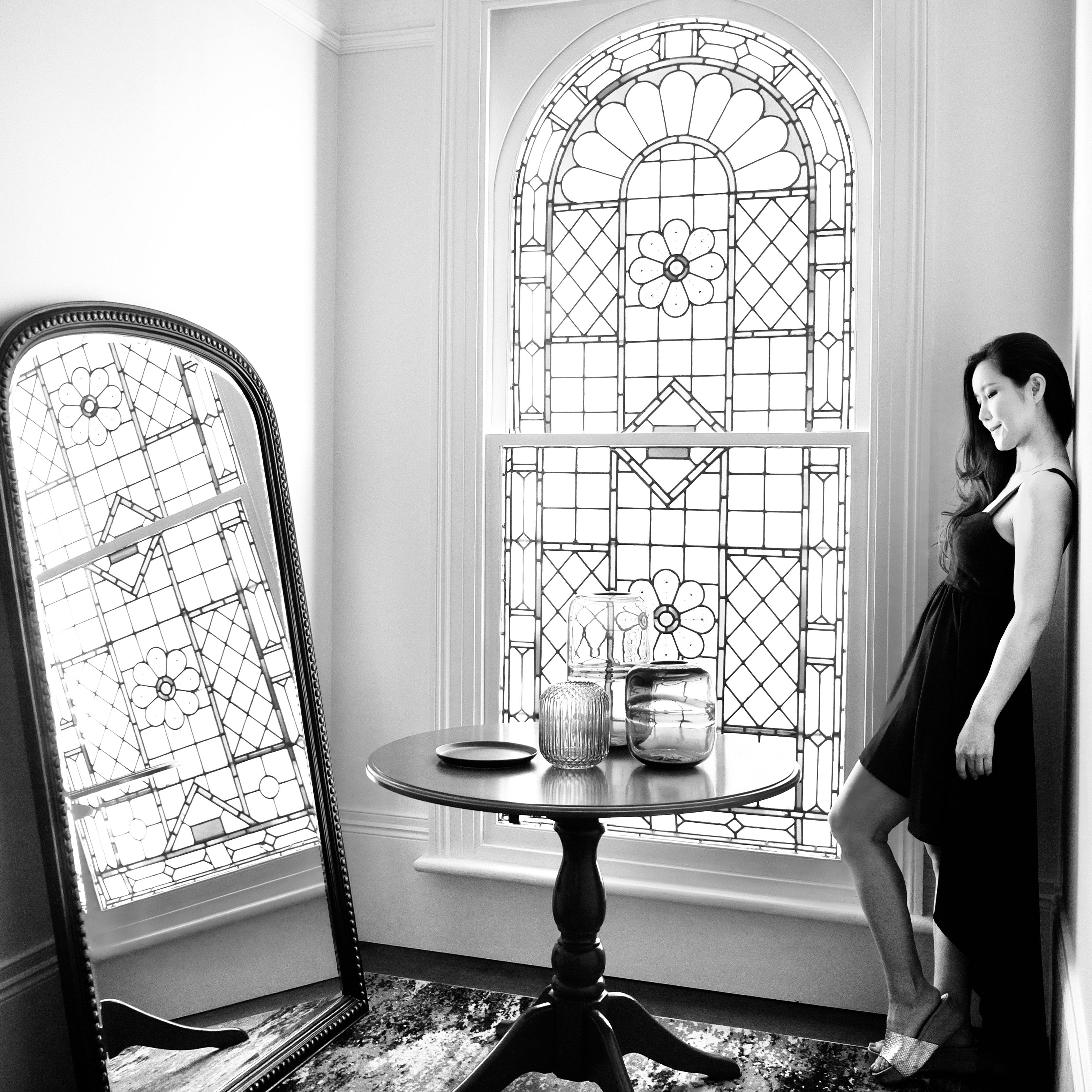 A stylish woman leans against a wall beside a stained glass window, reflected elegantly in a mirror.