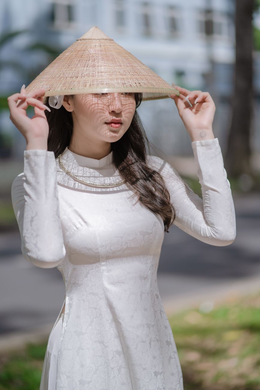 A woman wearing a traditional Vietnamese ao dai and conical hat poses outdoors in natural lighting.