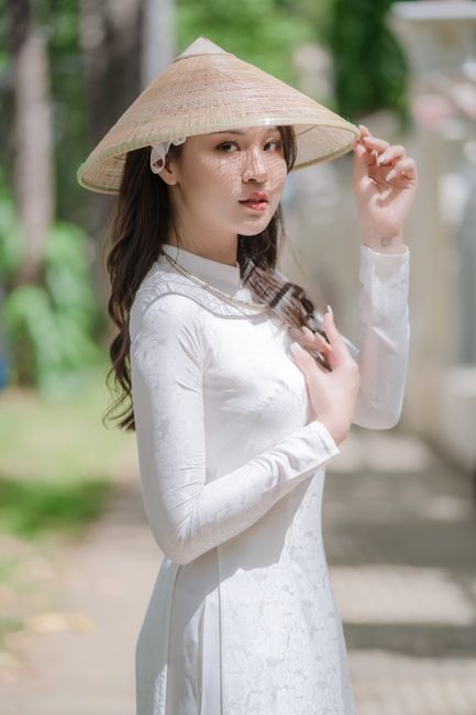 A woman in a white ao dai and conical hat poses gracefully outdoors, embodying traditional Vietnames