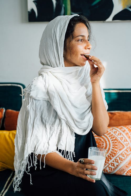 Free A Woman in White Headscarf Eating while Sitting on the Couch Stock Photo