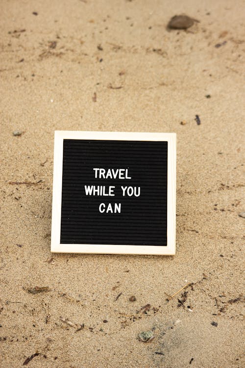 Board with Text 'Travel While You Can' on the Beach