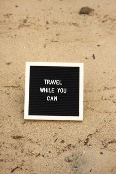 Inspirational message 'Travel while you can' on a signboard placed on beach sand, encouraging exploration.