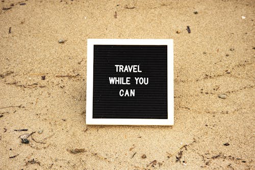 Board with Text 'Travel While You Can' on the Sand