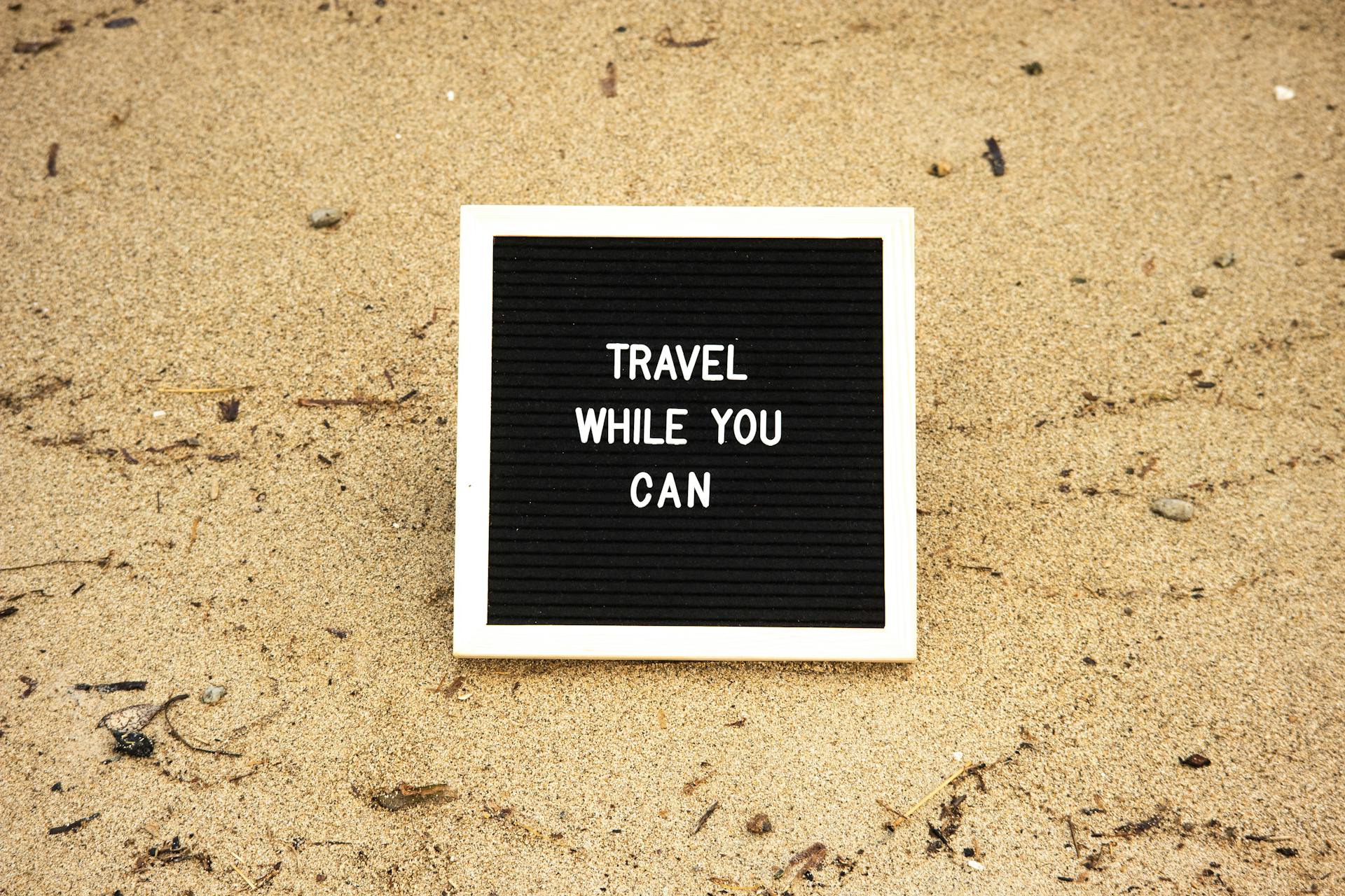 Quote 'Travel While You Can' on board in sand, inspiring travel and adventure.