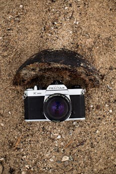 A classic Pentax camera placed on a sandy beach with an open coconut shell.