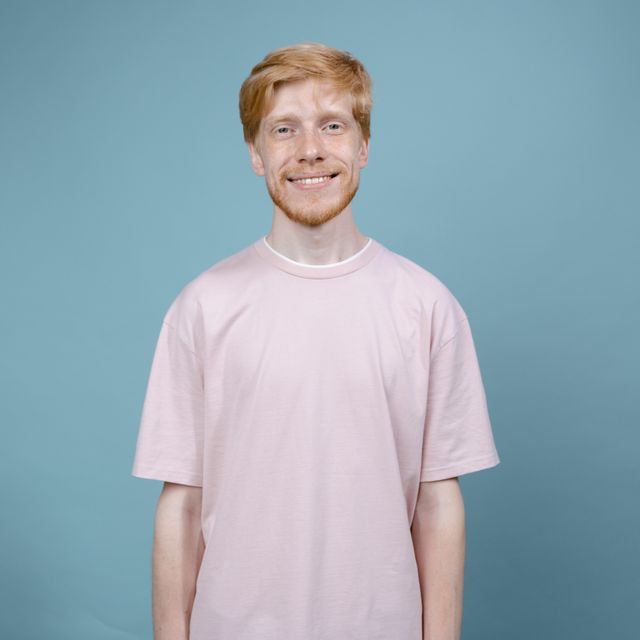 Junger Mann mit rotblonden Haaren und rosa T-Shirt lächelt vor blauem Hintergrund