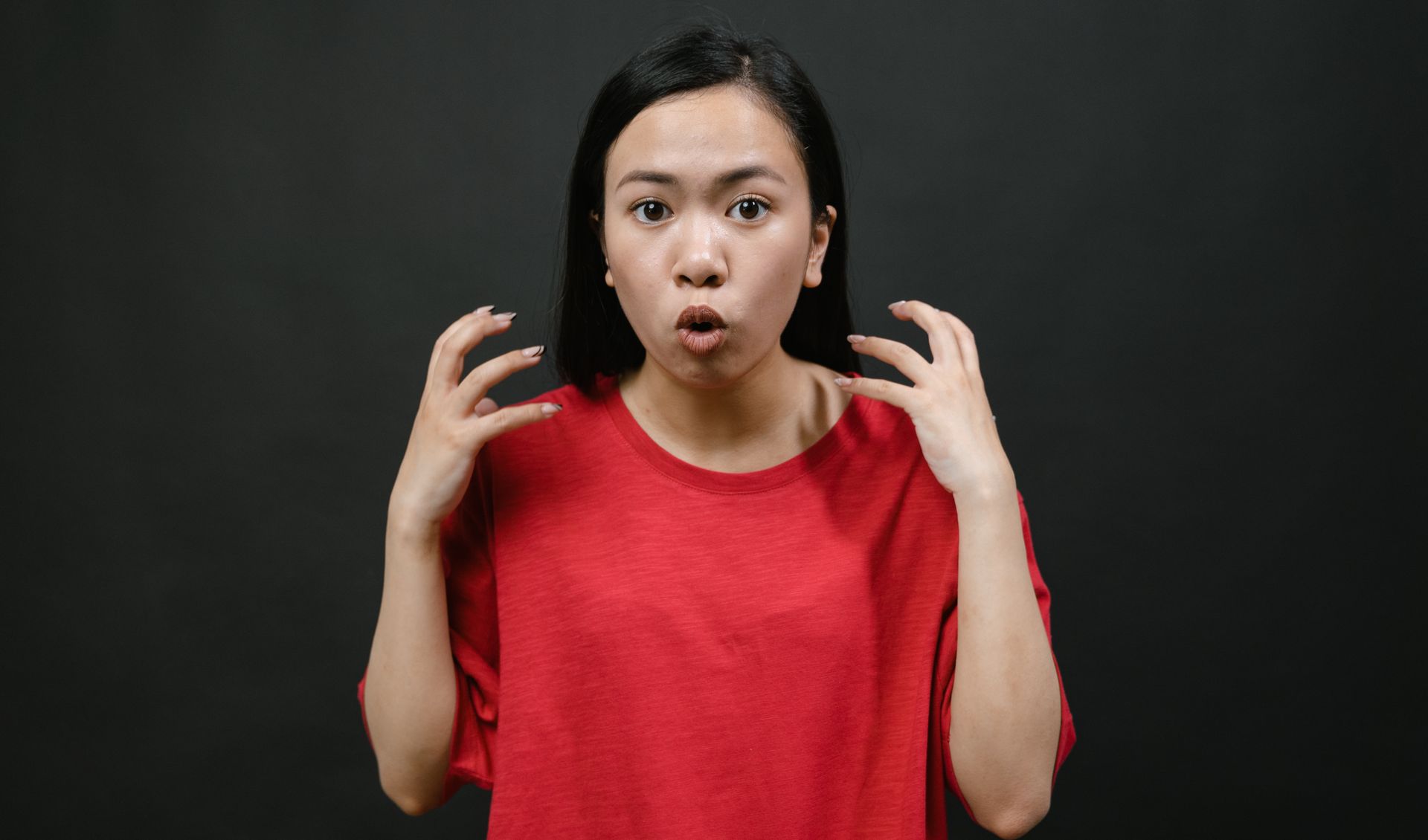 Asian woman in a red shirt expressing surprise with hands raised, on a dark background.