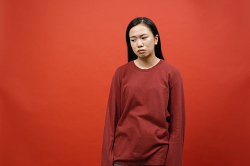 Free Portrait of an Asian woman standing against a deep red background, expressing sadness. Stock Photo