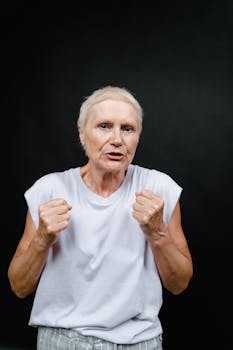 Senior woman exuding determination with clenched fists, wearing a white top against a black background.