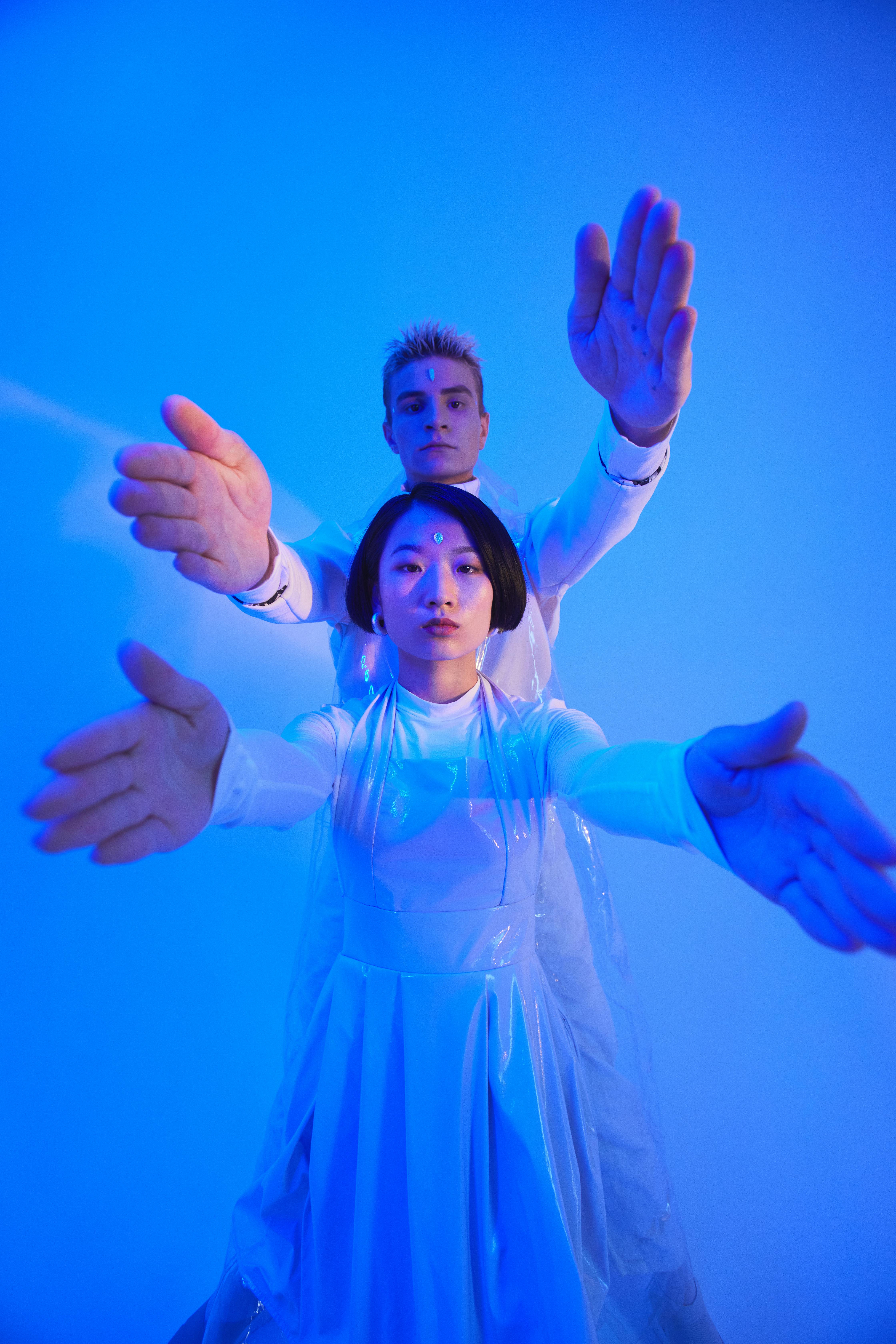 young man and woman in futuristic outfits standing in studio with blue lighting and holding their arms up