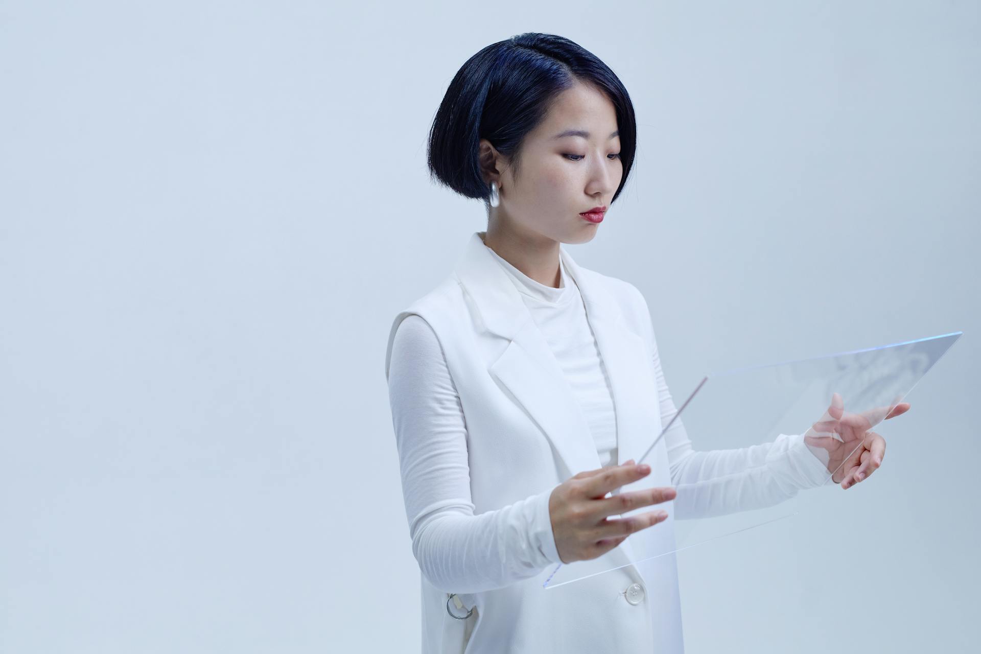 Asian woman in white blazer holding transparent glass interface on a plain background.