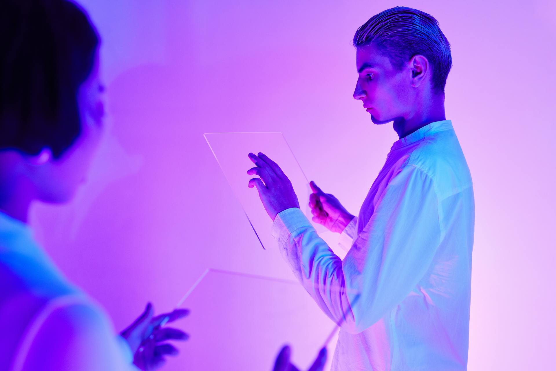 Two individuals engaging with futuristic transparent touch screens in a vibrant purple-lit studio.