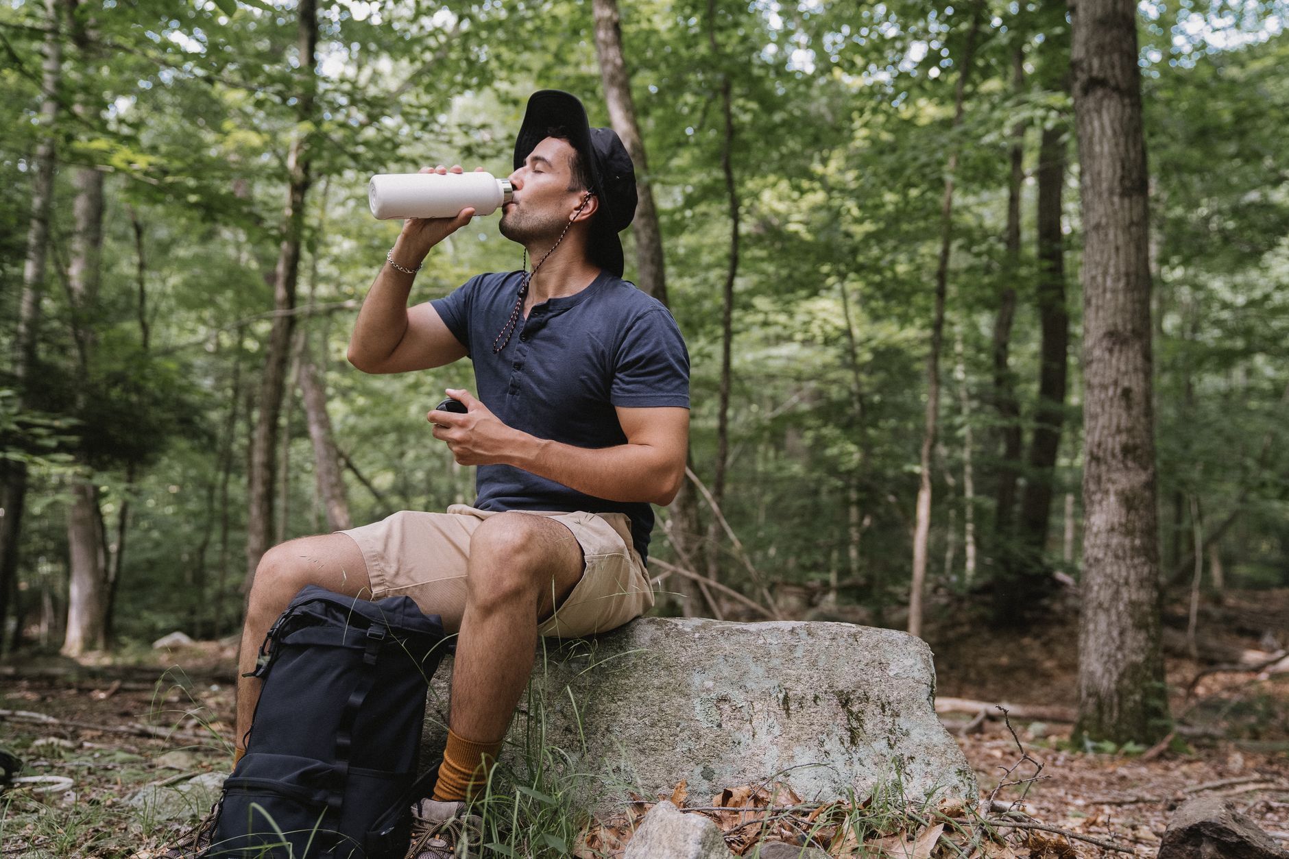 A man drinking water while resting on a rock in a lush forest during a hike.