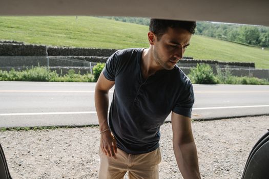 Young adult man standing by the open car on a sunny rural roadside.