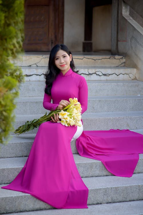 Free A Pretty Woman in Pink Dress Holding Yellow Flowers while Sitting on a Concrete Staircase Stock Photo