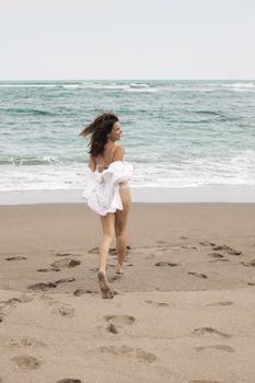 A carefree young woman runs along a serene beach with waves crashing nearby.