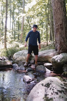A man wearing a hat steps across rocks in a serene forest stream, surrounded by lush greenery.