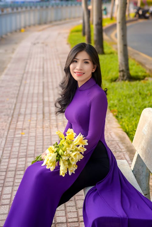Free A Pretty Woman in Purple Dress Sitting while Holding Yellow Flowers Stock Photo