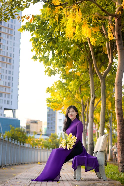 Free A Pretty Woman in Purple Dress Sitting while Holding Yellow Flowers Stock Photo