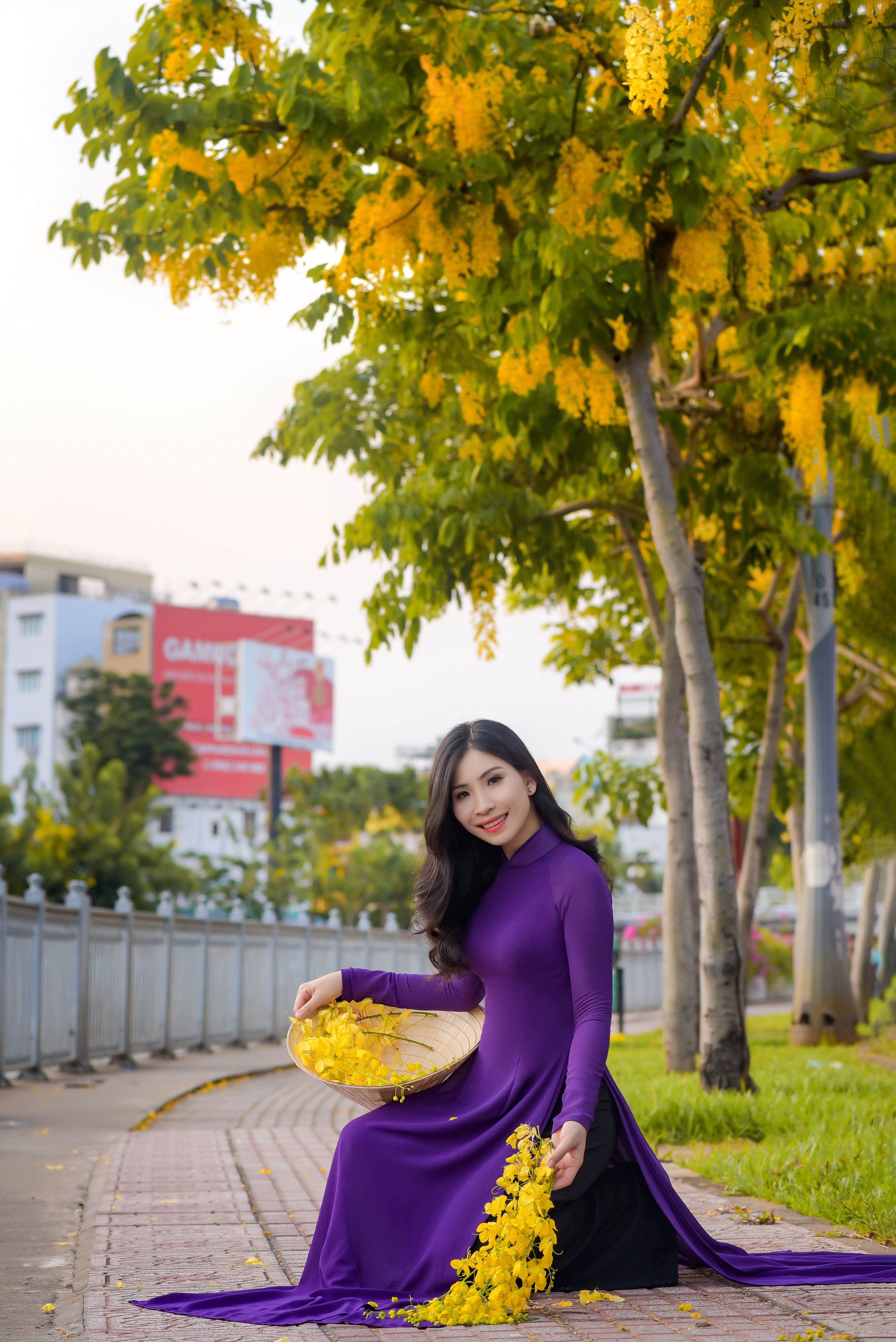 Elegant Asian woman in purple Ao Dai posing with yellow flowers outdoors.