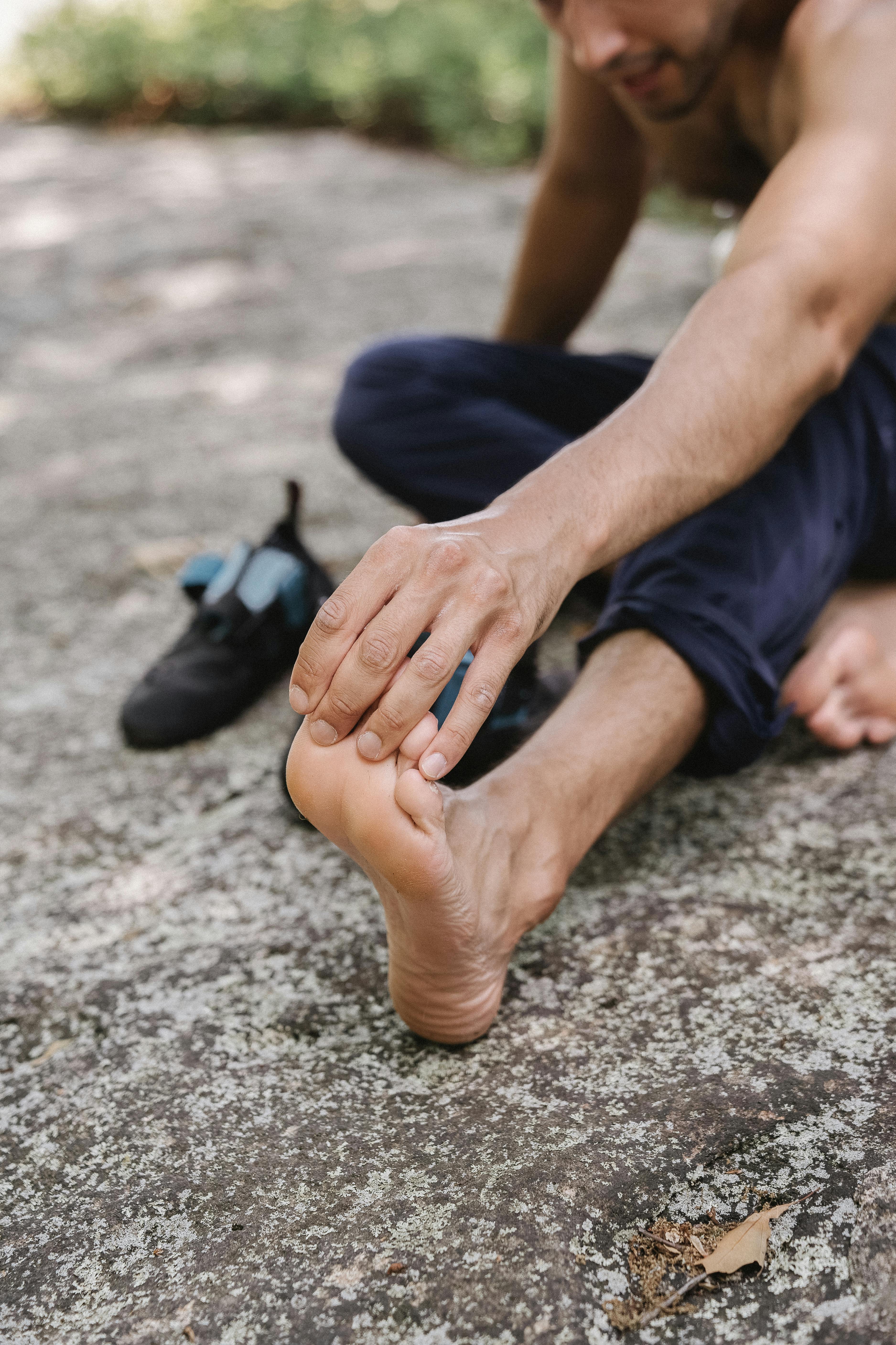 A shirtless man sits on the ground stretching his foot on a warm day.