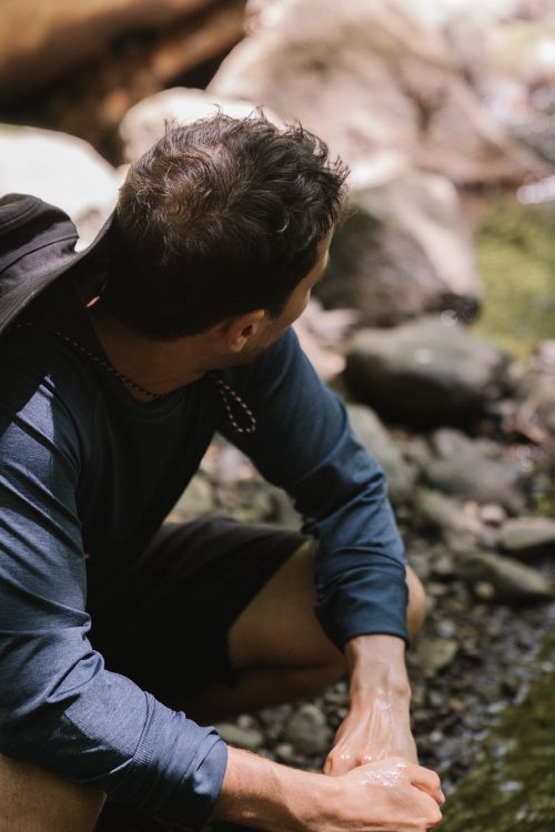 Free A man with a backpack crouches beside a serene stream, enjoying nature's tranquility. Stock Photo