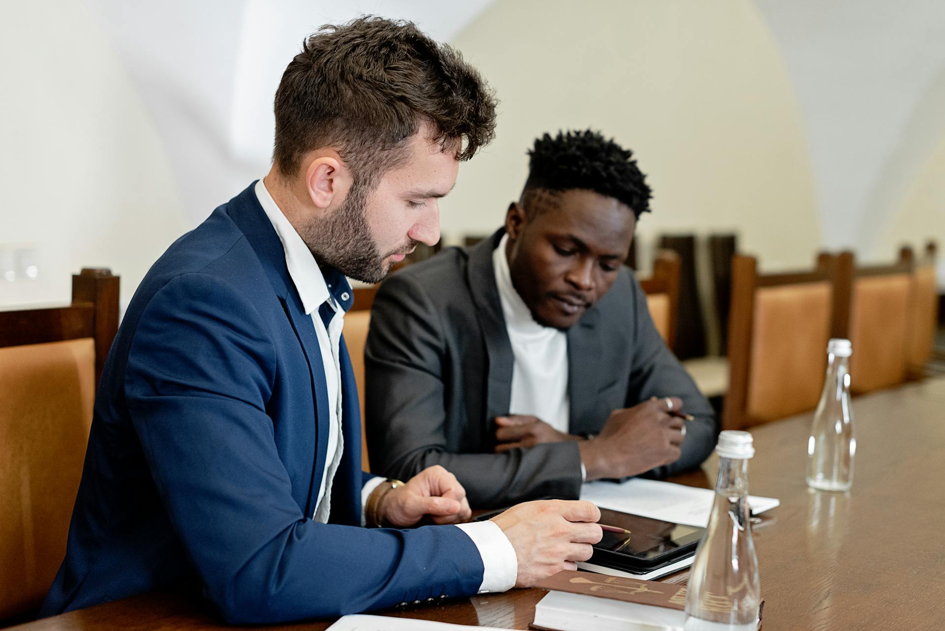 Two businessmen in suits discussing a project using a digital tablet during a business meeting indoors.