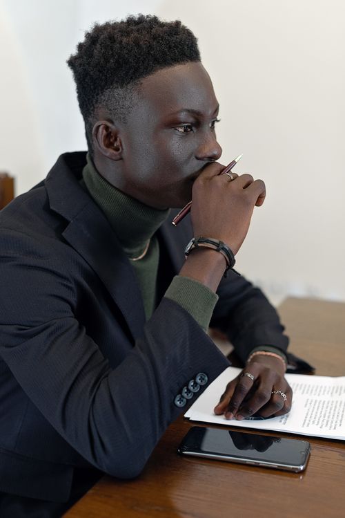 Free Young man in blazer sitting at table, holding pen in thoughtful pose. Stock Photo