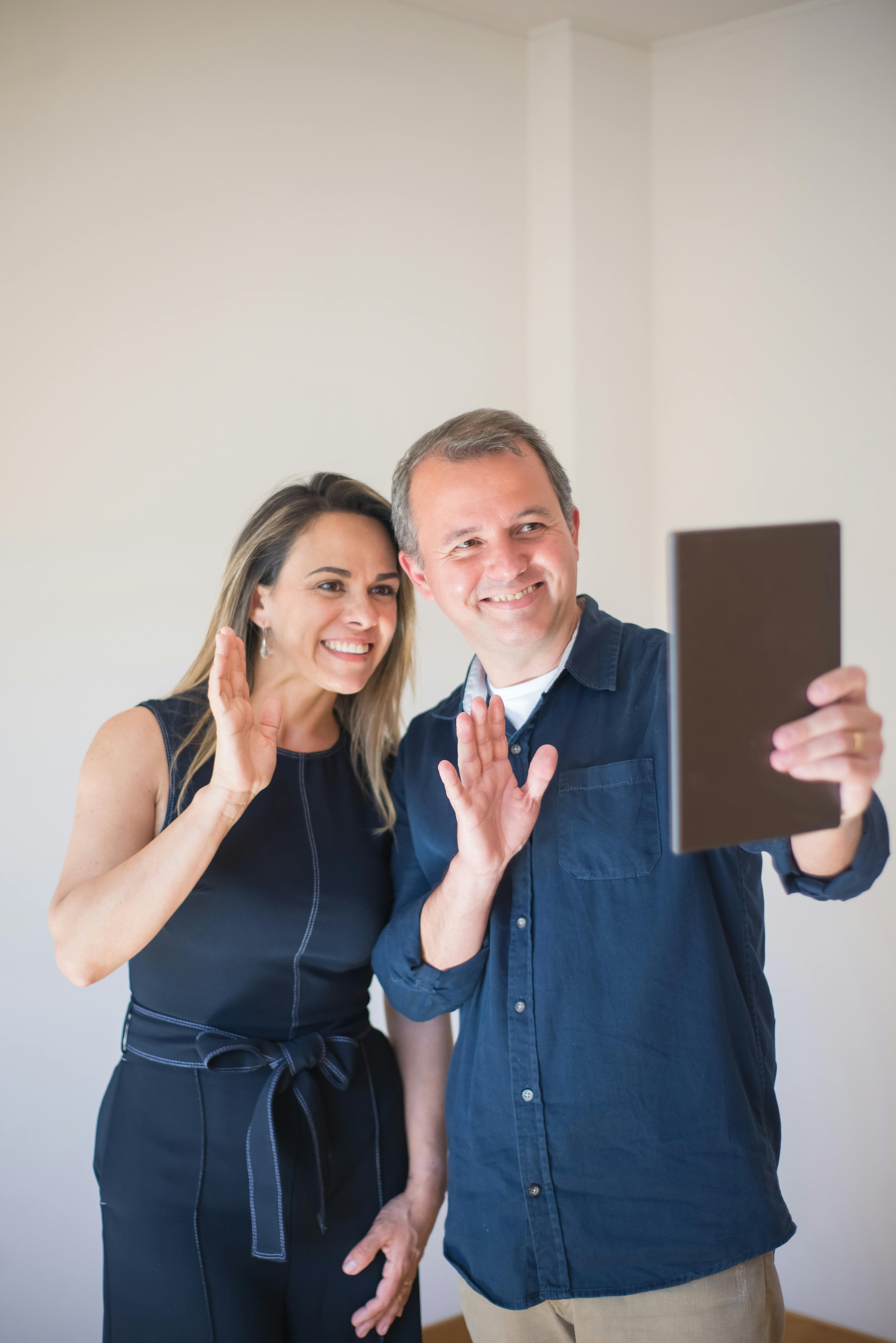 A joyful couple making a videocall from their bright home interior, showing a welcoming gesture.
