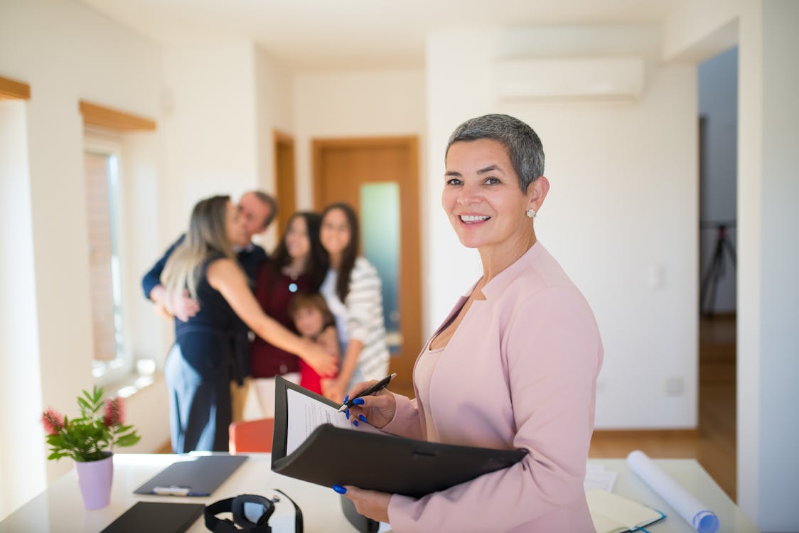 Real estate agent with a clipboard smiling at the camera, with a family embracing in their new home in the background, signifying a successful property purchase.