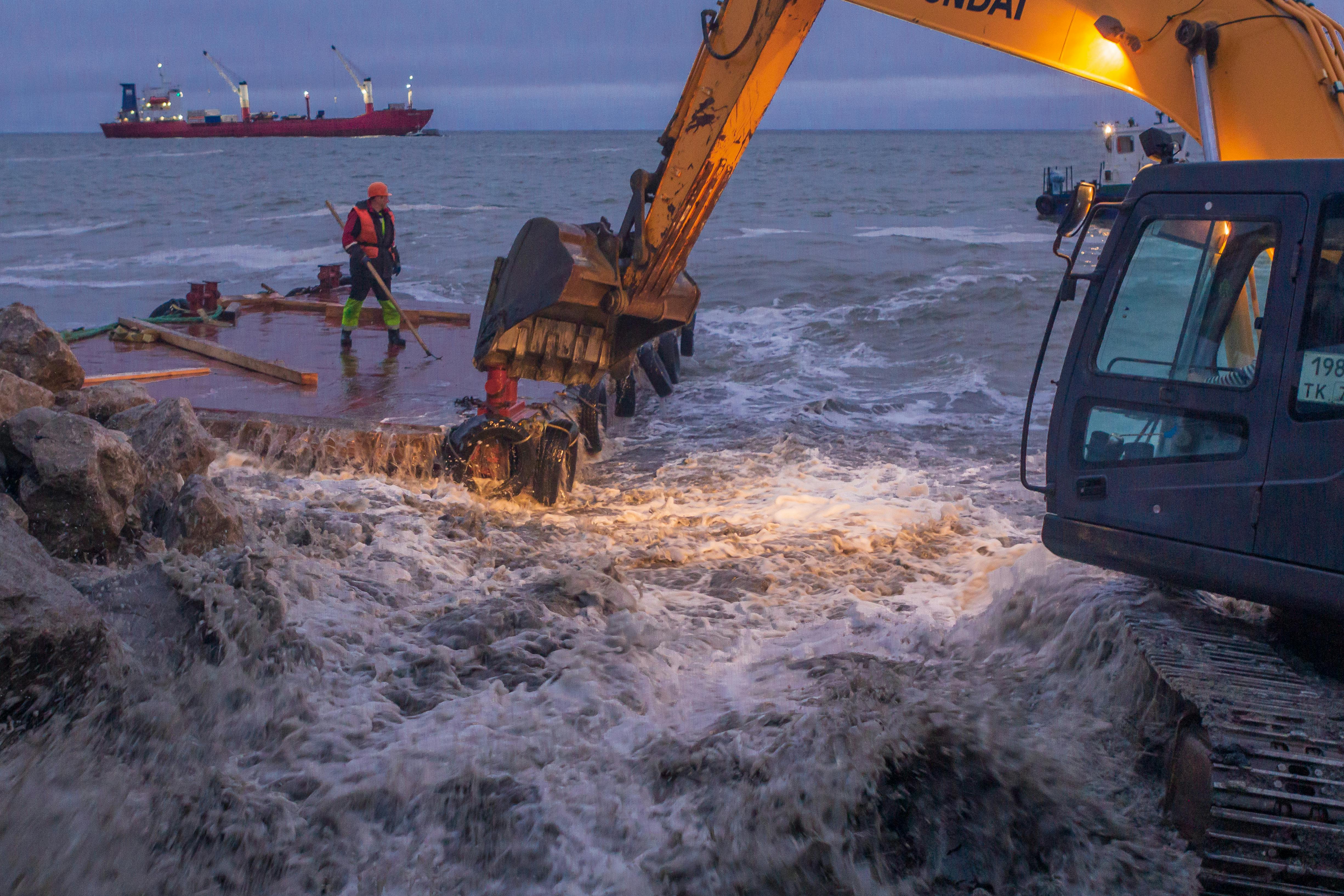 An excavator operates near crashing waves with a container ship in the background.