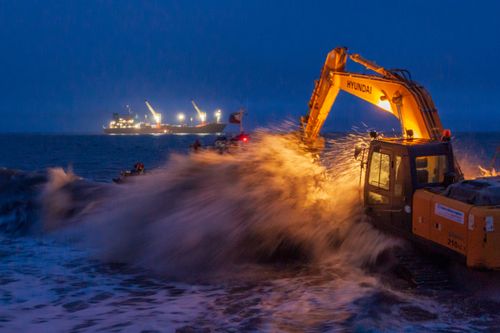 Free An excavator operates by the sea at night, splashing waves against its arm under vivid ship lights. Stock Photo