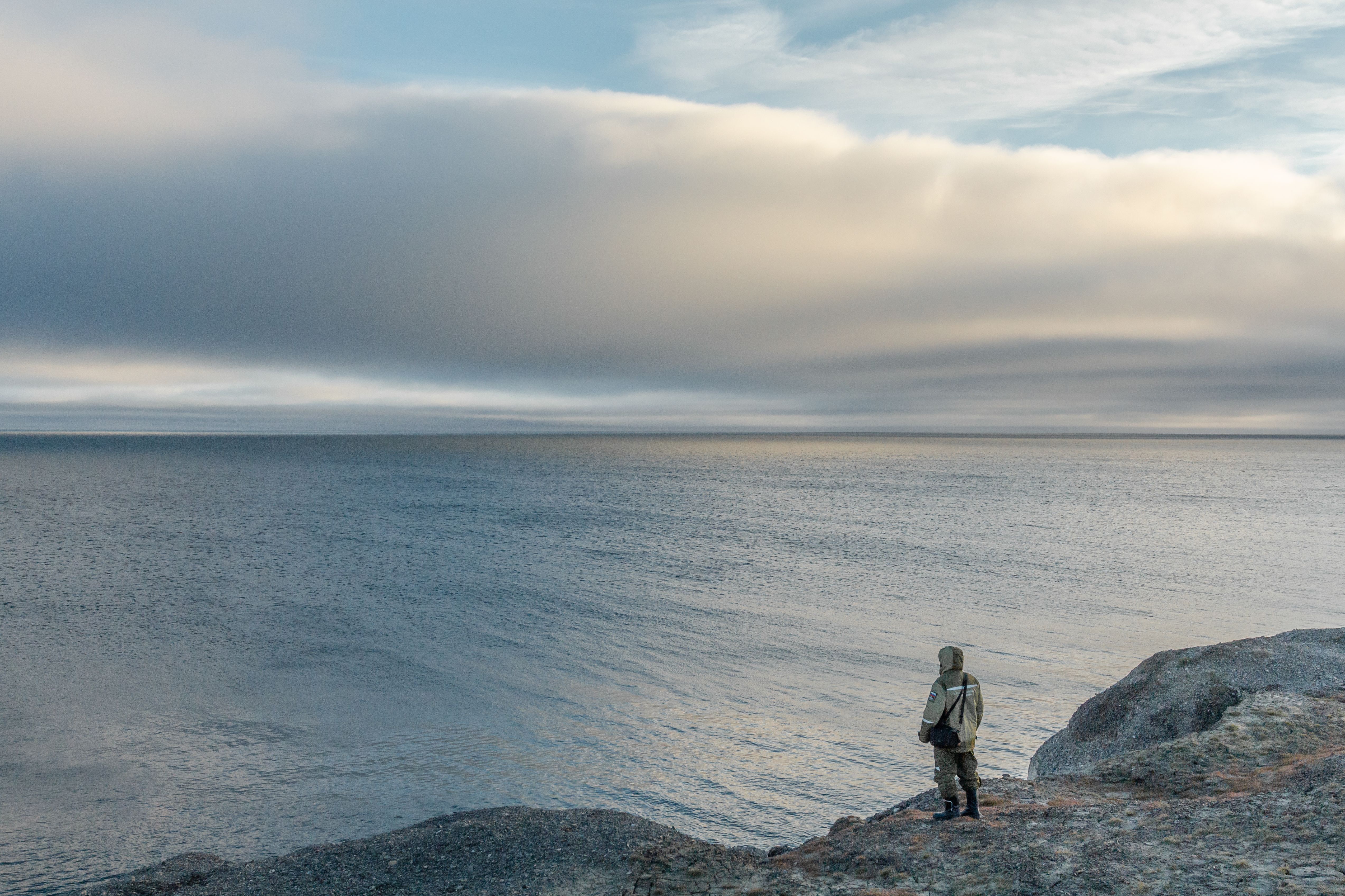 A lone person stands on rocky shore, gazing at the tranquil sea under a cloudy sky.