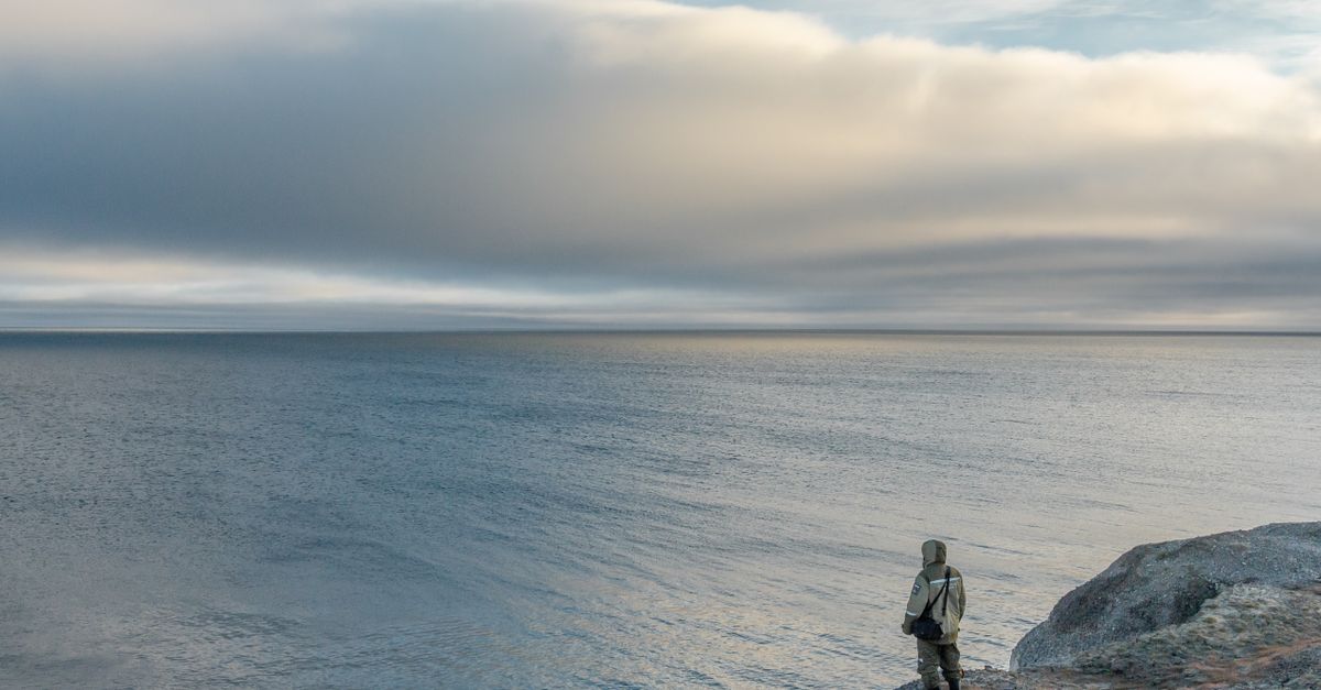 A lone person stands on rocky shore, gazing at the tranquil sea under a cloudy sky.