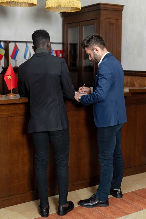 Free Two men in suits signing documents at a hotel reception desk, indicating partnership and professionalism. Stock Photo