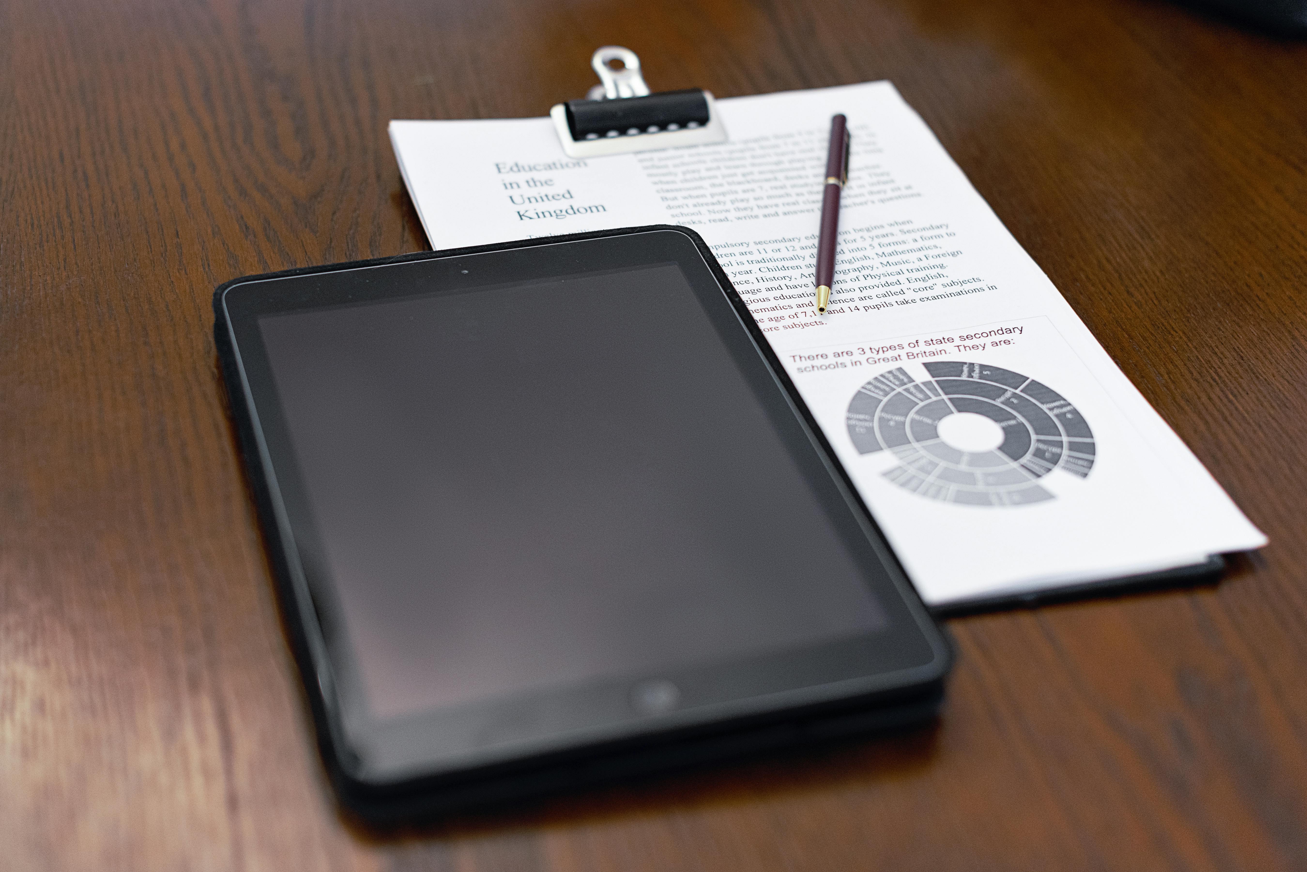 A tablet and educational documents on a wooden table with a pen.