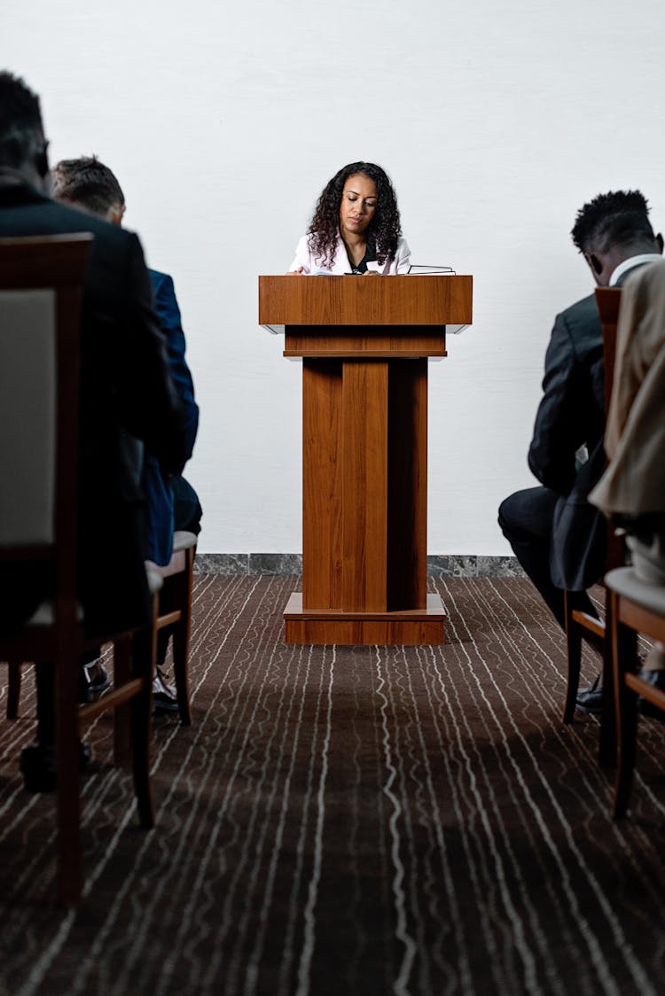 A Woman Standing At A Podium In Front Of People Sitting On Chairs